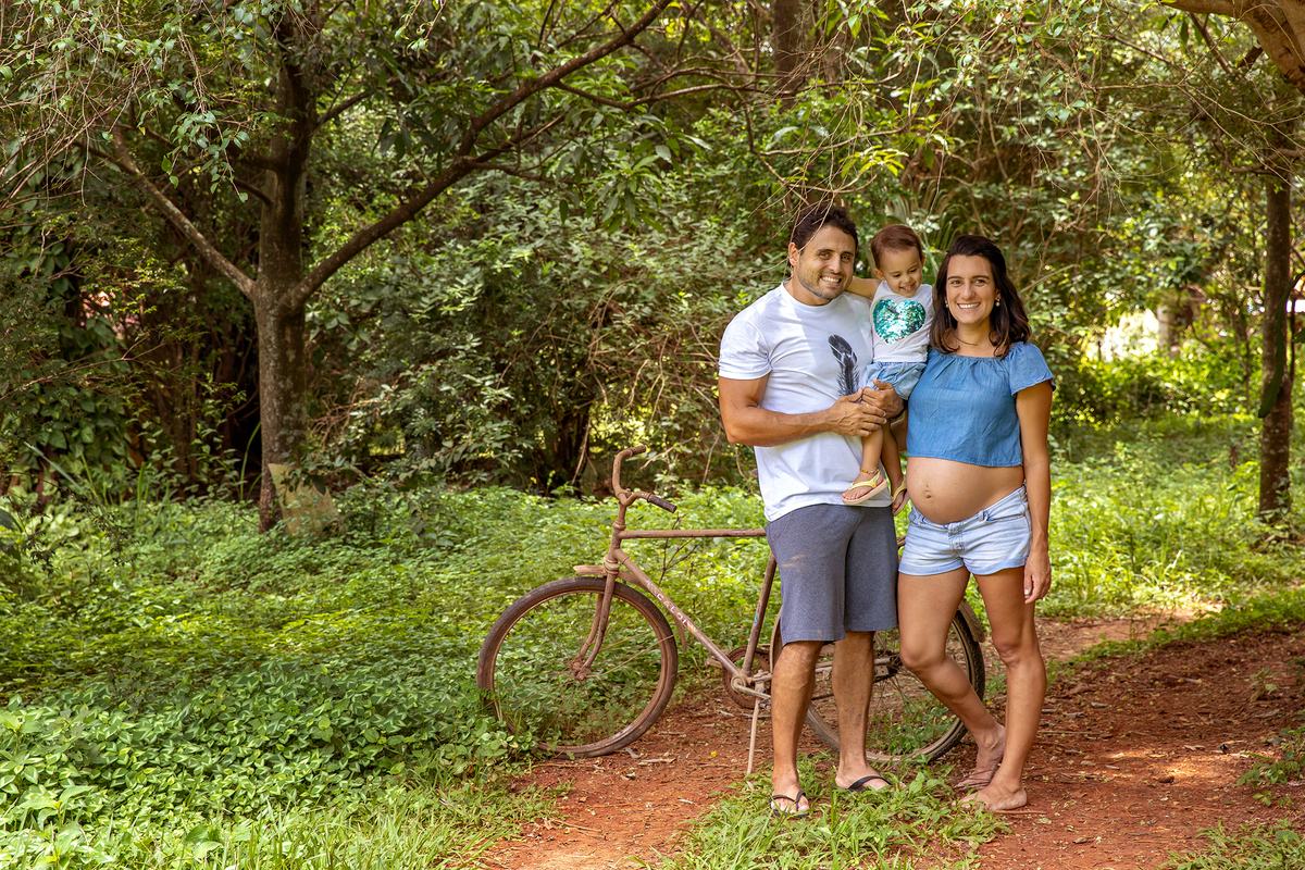 Mulher GRÁVIDA com a BARRIGA de fora, posando com o MARIDO e a FILHA em ENSAIO GESTANTE em um SÍTIO, em click do FOTÓGRAFO de GESTANTE.