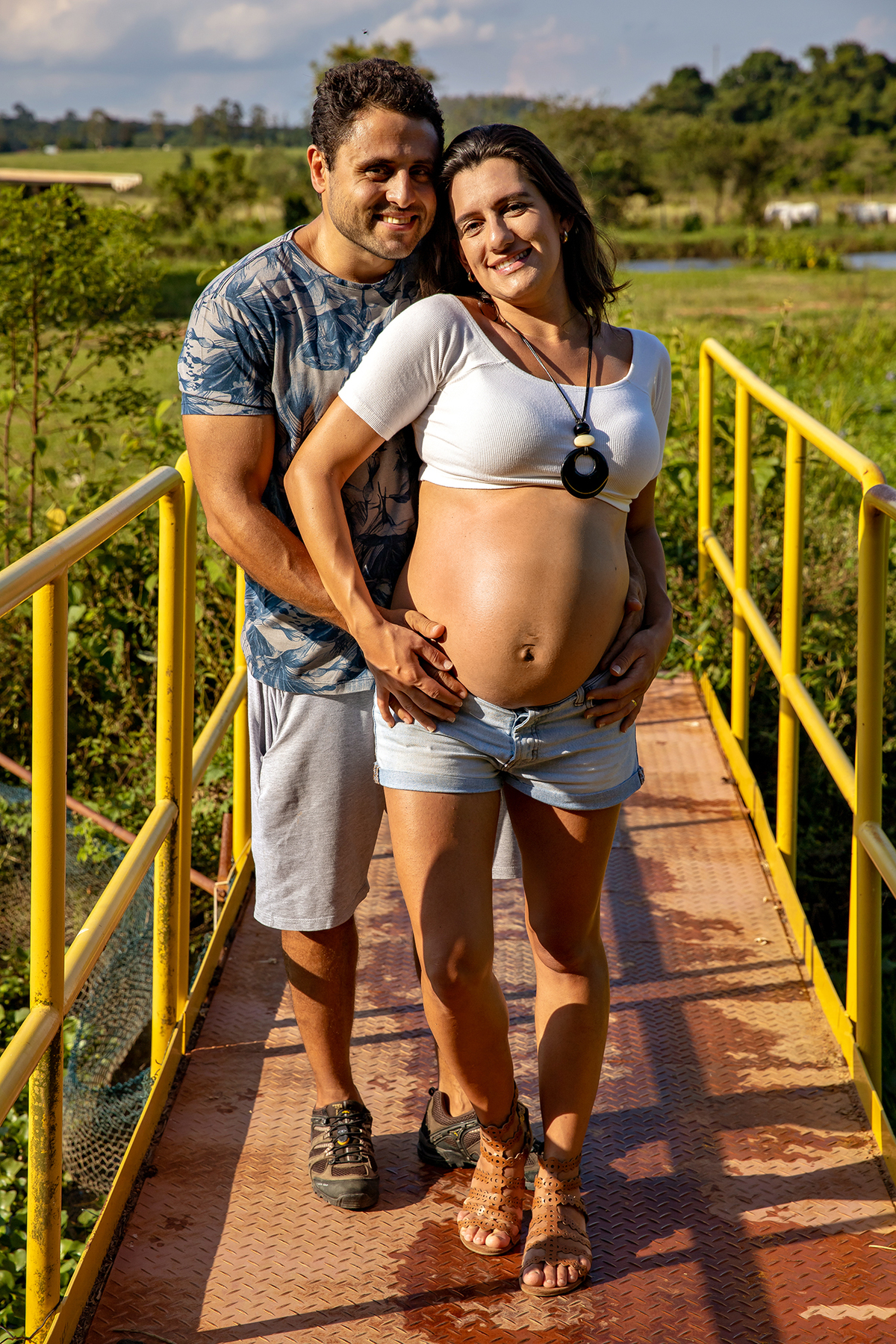Mulher GRÁVIDA com a BARRIGA de fora, posando com o MARIDO em ENSAIO GESTANTE em um SÍTIO, em click do FOTÓGRAFO de GESTANTE.