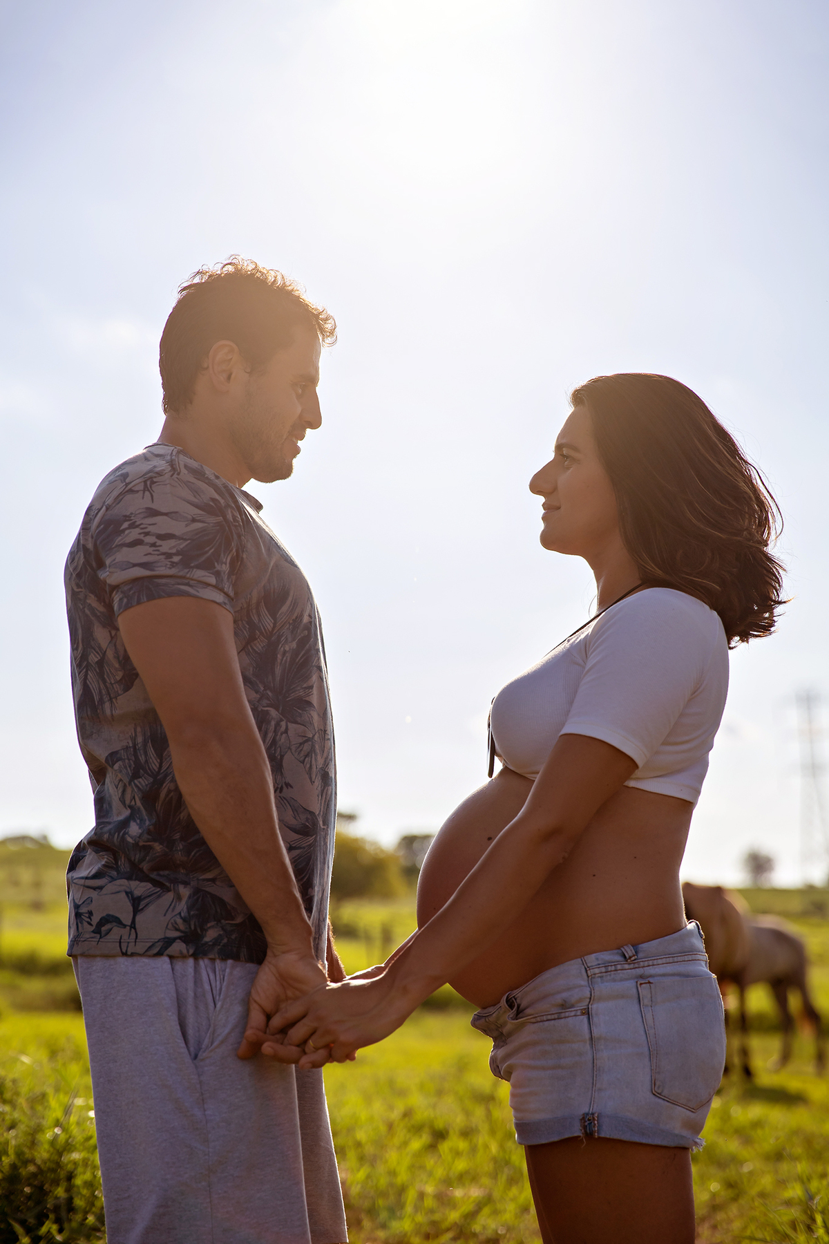 Mulher GRÁVIDA com a BARRIGA de fora, posando com o MARIDO em ENSAIO GESTANTE em um SÍTIO, em click do FOTÓGRAFO de GESTANTE.