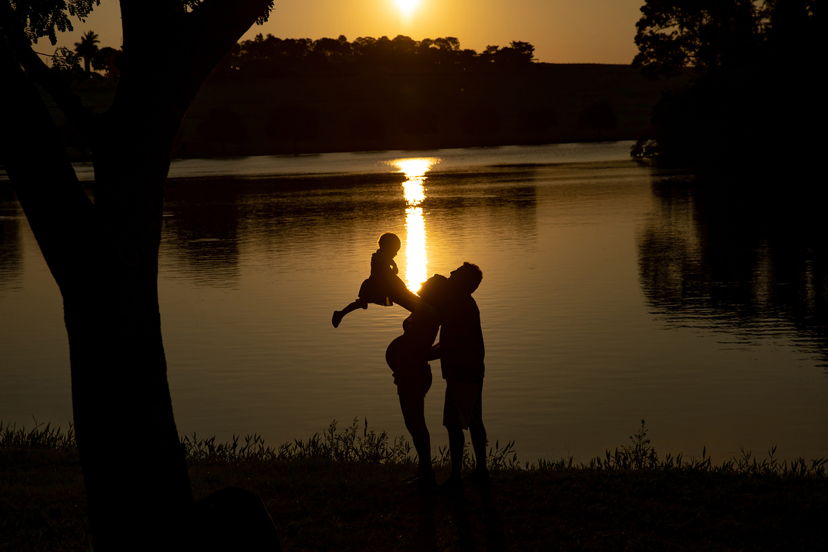 Mulher GRÁVIDA com a BARRIGA de fora, posando com o MARIDO e a FILHA em ENSAIO GESTANTE em um SÍTIO no POR do SOL, em click do FOTÓGRAFO de GESTANTE.