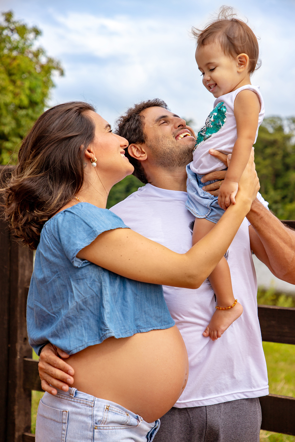Mulher GRÁVIDA com a BARRIGA de fora, posando com o MARIDO e a FILHA em ENSAIO GESTANTE em um SÍTIO, em click do FOTÓGRAFO de GESTANTE.
