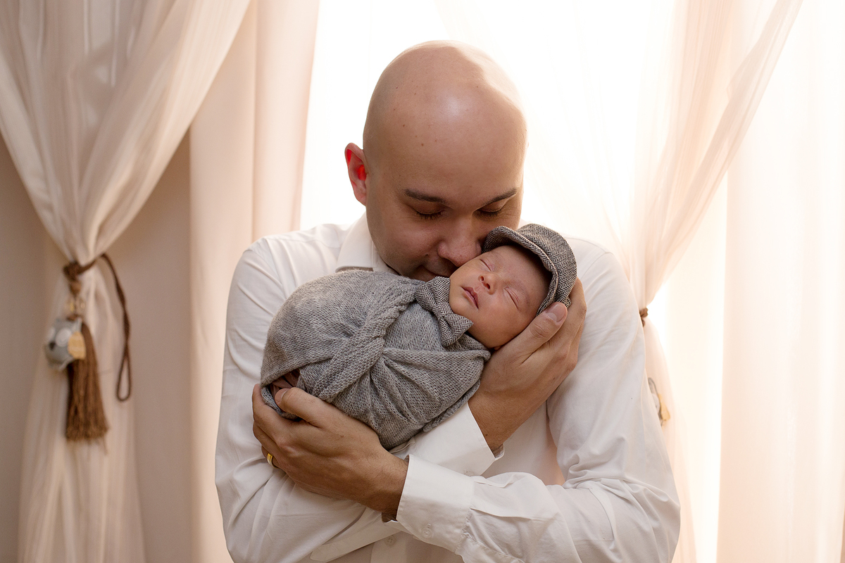 Ensaio Newborn em casa foto n o quartinho do bebê foto com pai dando cheirinho no bebê bebê recém nascido enroladinha em tons de cinza com boina e gravatinha borboleta
