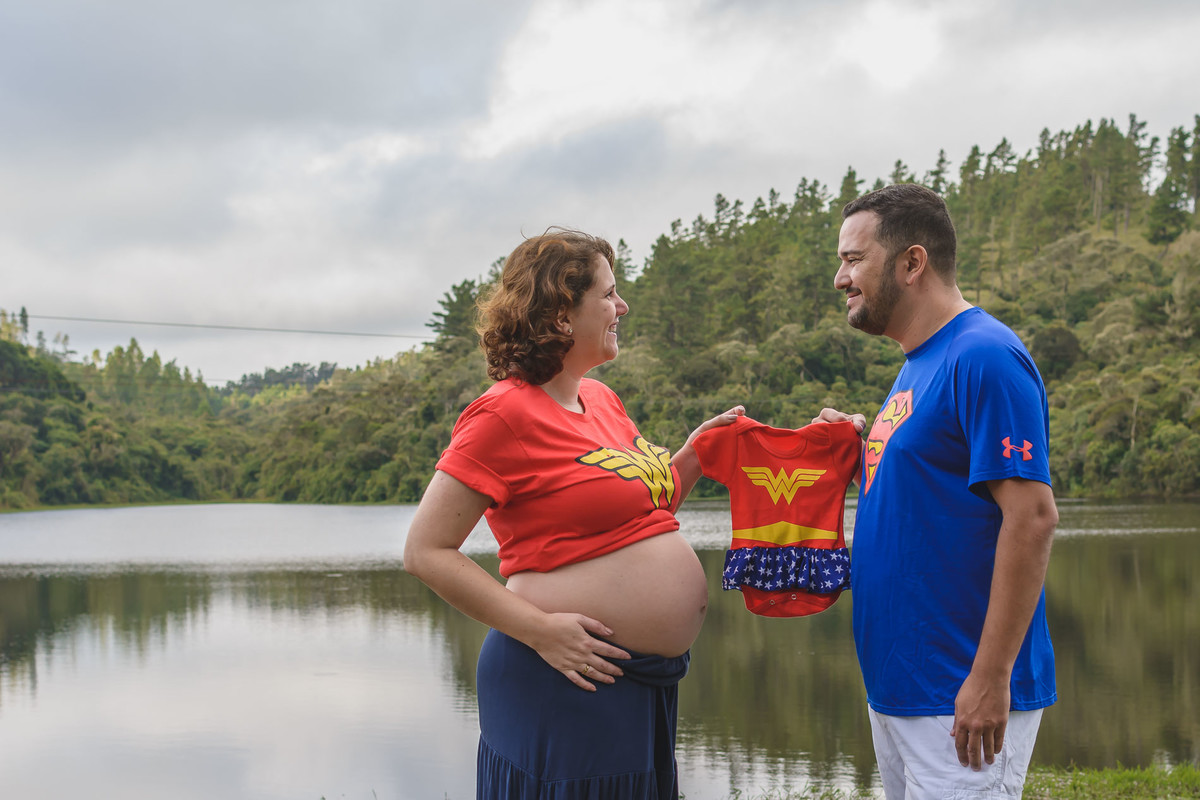 Fotografia do casal segurando a roupinha de Mulher Maravilha do bebê - ensaio de gestante temático em Campos do Jordão - Cia P&B - Fotografia