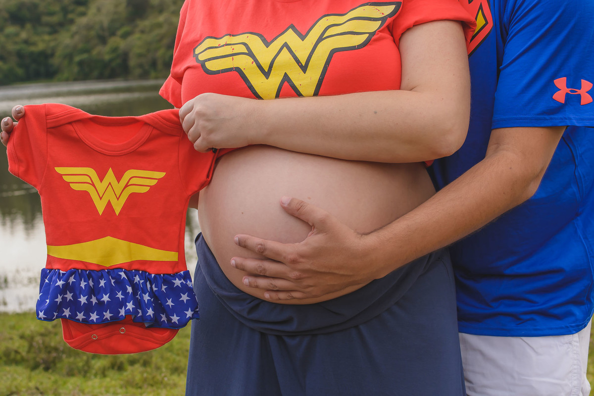 Fotografia da mão na barriga da gestante com a roupinha do bebê de Mulher Maravilha sendo mostrada - ensaio de gestante temático em Campos do Jordão - Cia P&B - Fotografia