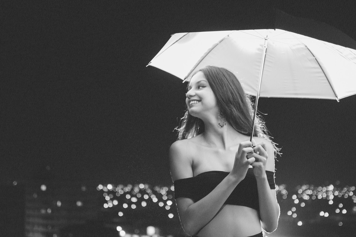Fotografia de debutante sorrindo com a sombrinha e efeito de chuva com luzes ao fundo - Cia P&B - Fotografia - Praia Grande