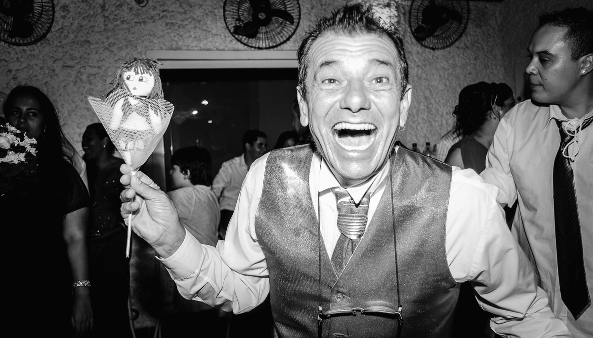 Fotografia do pai da noiva sorrindo com o santo antônio do buquê  durante a festa de casamento no Buffet Laranja em Mongaguá - cia P&B - Fotografia