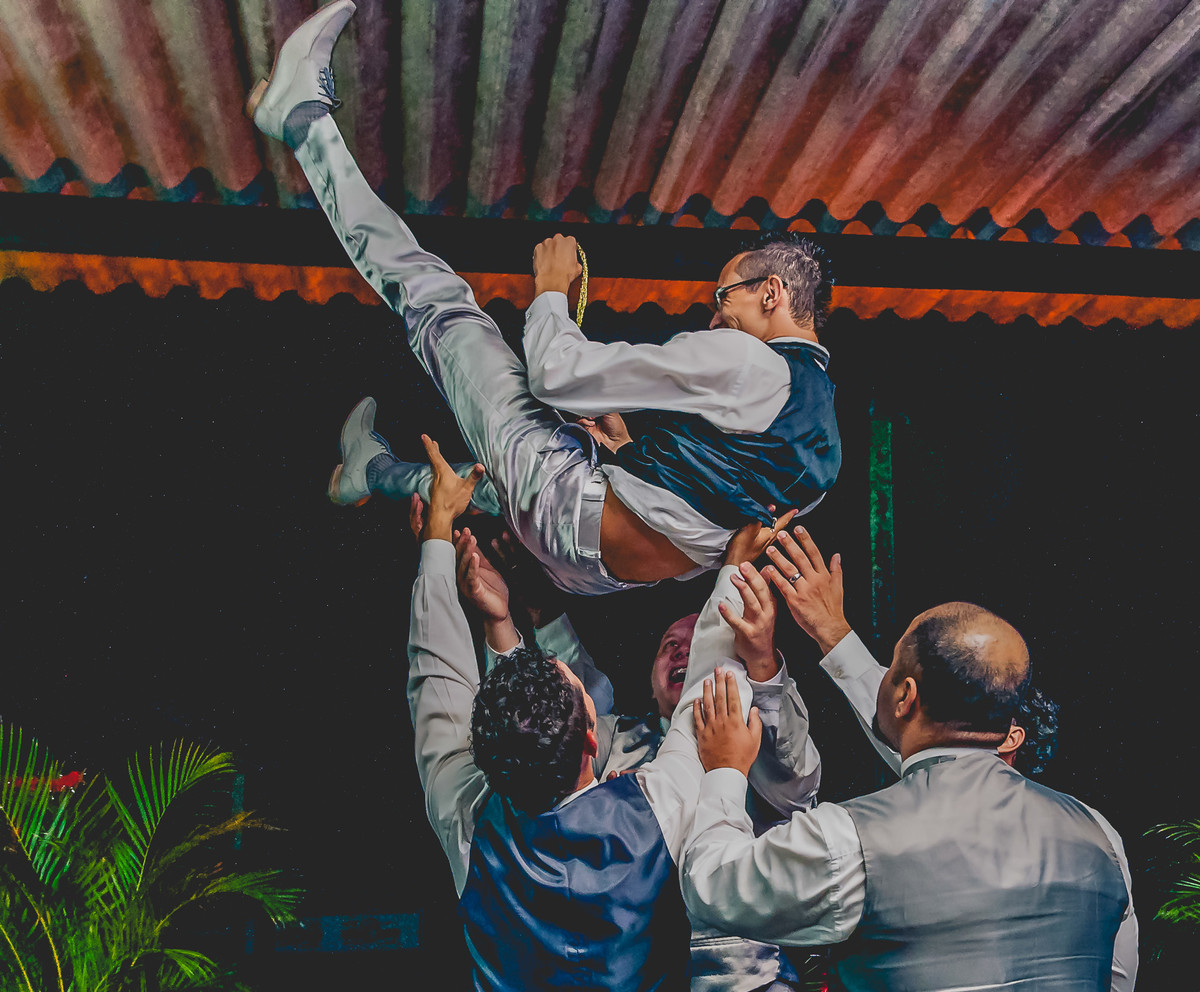 Fotografia do noivo sendo jogado para cima durante a festa de casamento na Assebam em Mongaguá - Cia P&B - Fotografia 