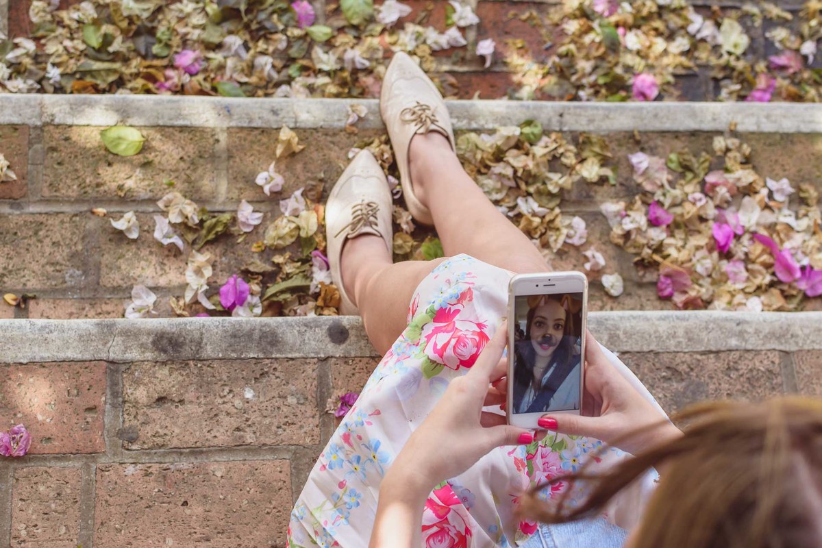 Fotografia da blogueira Fofuras da Ka mostrando detalhes das flores no chão e ela segurando seu celular durante ensaio fotográfico em Itanhaém - Cia P&B - Fotografia 