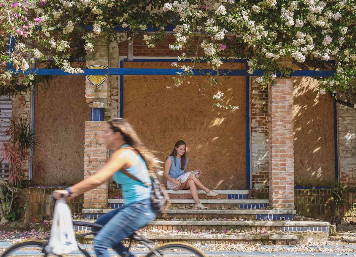 Fotografia da blogueira Fofuras da Ka sentada na escada com flores e mulher de bicicleta passando durante o ensaio fotográfico em Itanhaém - Cia P&B - Fotografia 