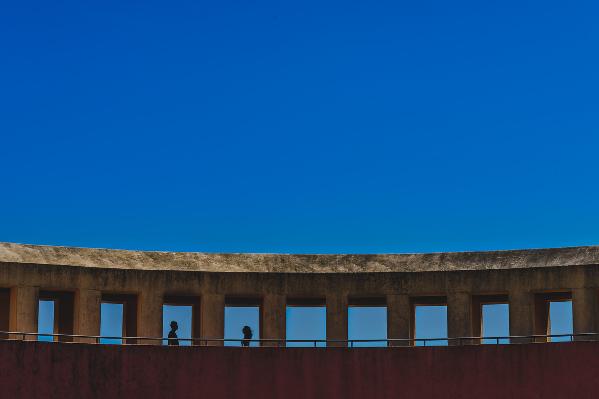 Foto dos Noivos de frente um para o outro em silhueta, com céu azul ao fundo e a estrutura principal da entrada do Parque Tanguá no ensaio pré wedding - Curitiba - fotografia Cia P&B Produções 