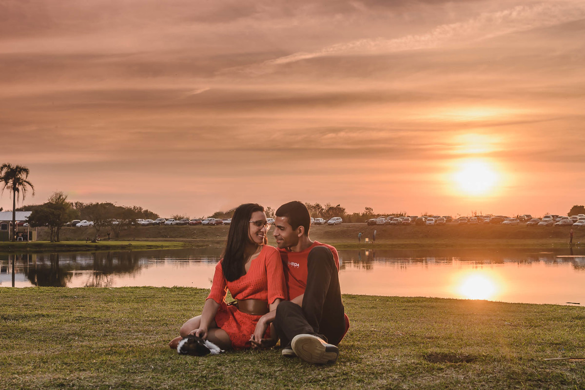 Foto dos Noivos sentados no gramado se olhando e sorrindo com o cachorrinho filhote deles com pôr do sol ao fundo no parque  São José dos Pinhais no ensaio pré wedding - PR - fotografia Cia P&B Produções 