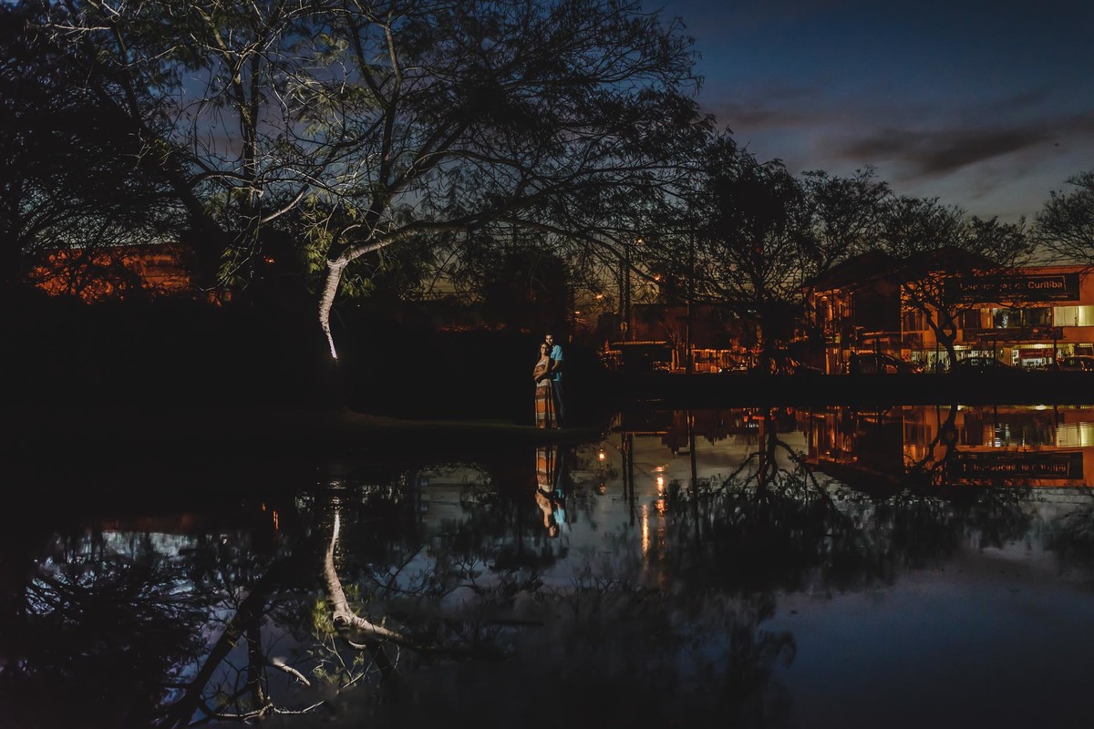Foto minimalista dos noivos abraçados com reflexo no lago das árvores, dos noivos e das casas iluminadas ao fundo no ensaio pré wedding - Jardim Botânico - Curitiba - fotografia Cia P&B Produções 
