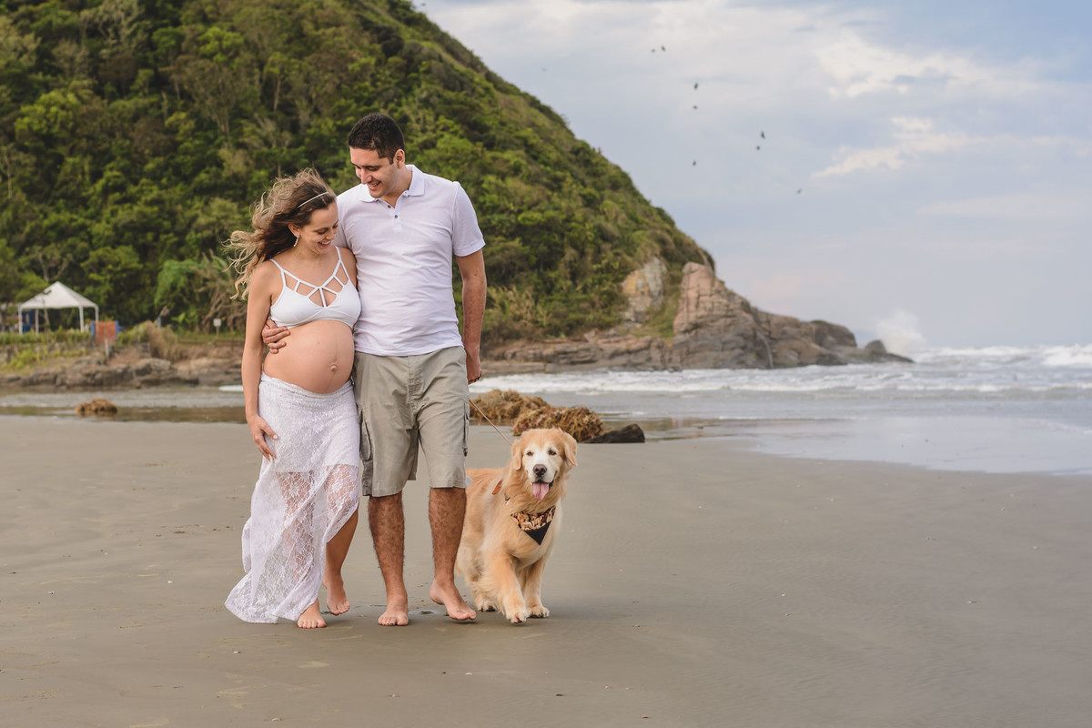 Foto da gestante e marido caminhando na praia com o cachorro do casal. Ensaio de gestante feito pela Cia P&B Produções Itanhaém - SP