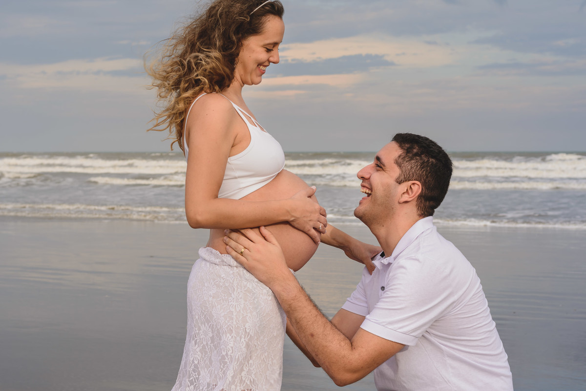 Foto da gestante com o marido sorrindo na praia. Ensaio de gestante feito pela Cia P&B Produções Itanhaém - SP