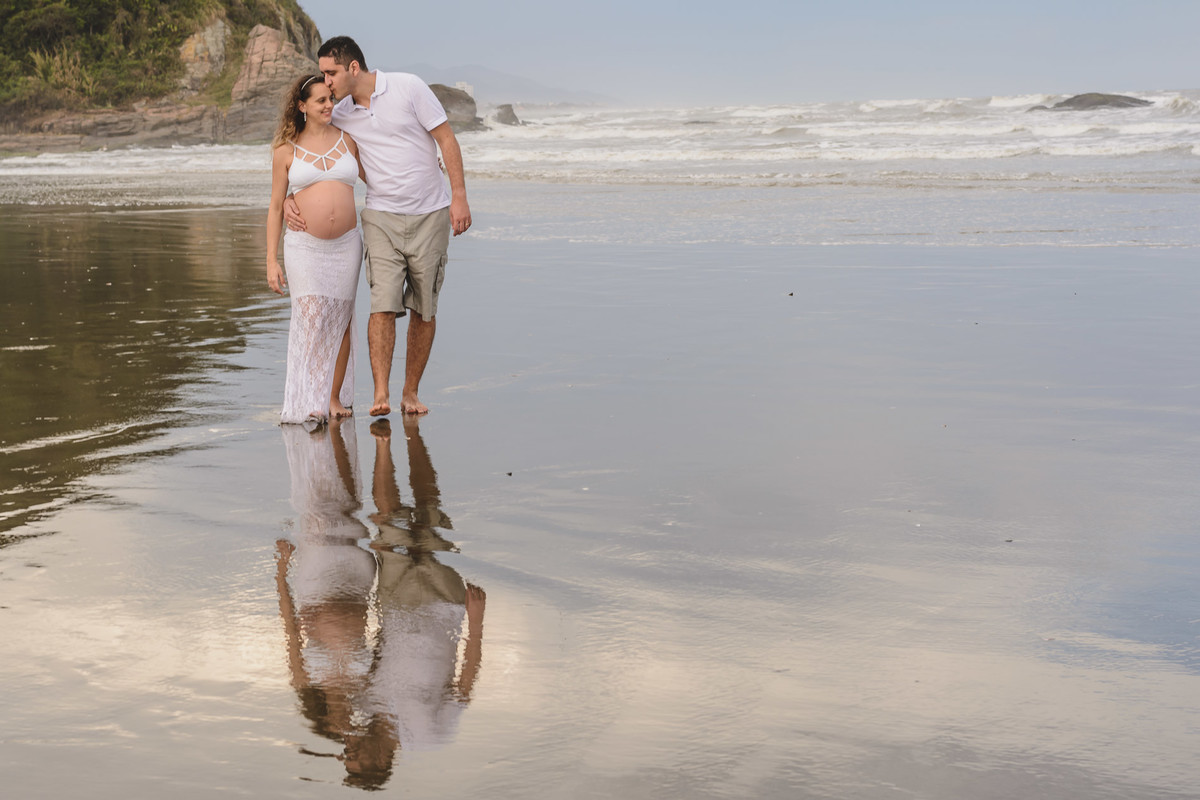 Foto da gestante com o marido com reflexo no mar na praia. Ensaio de gestante feito pela Cia P&B Produções Itanhaém - SP
