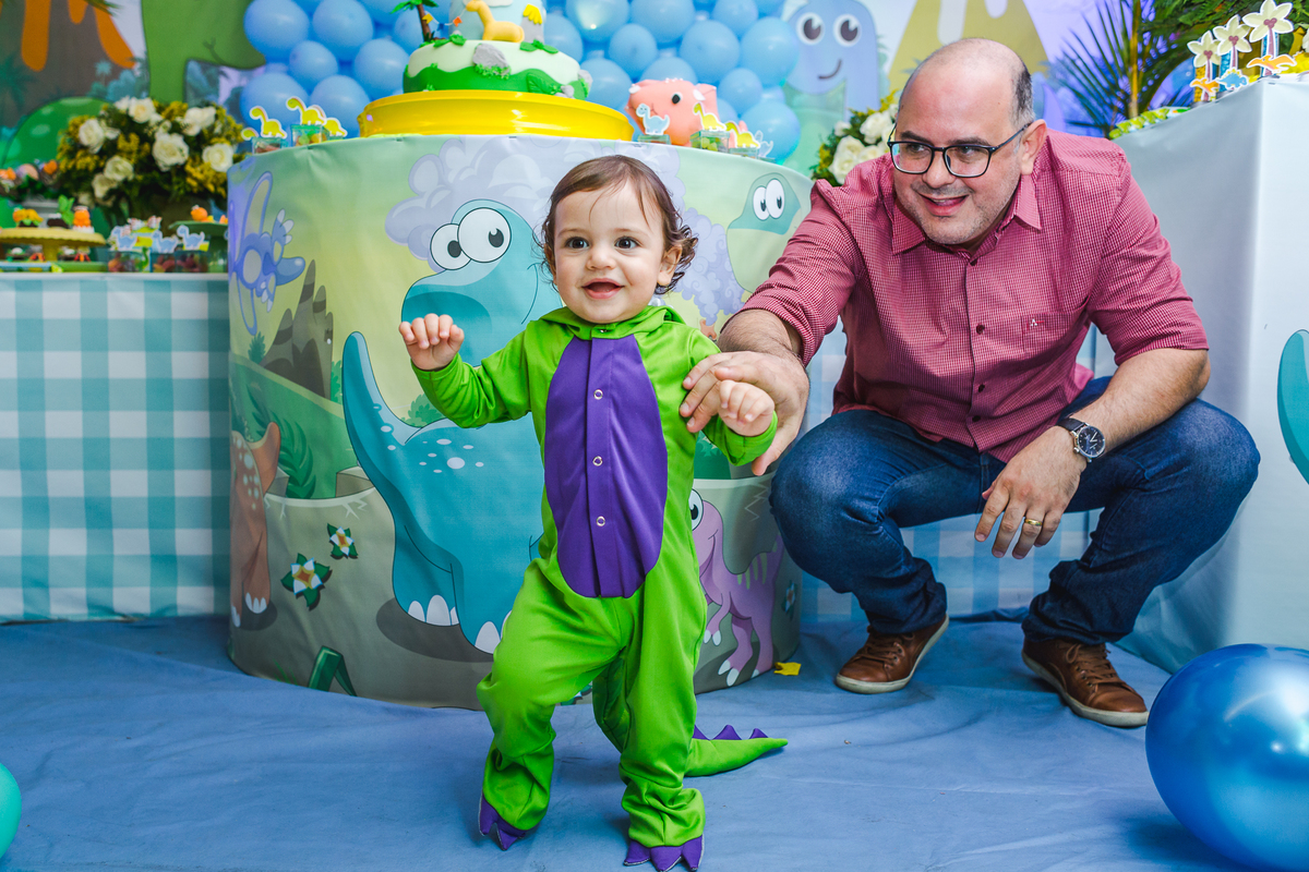 bebe com roupa de dinossauro andando sozinho, e pai atras rindo e observando