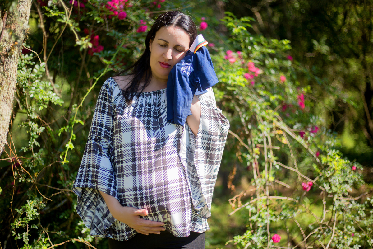a gestante segura a roupinha do bebê próximo ao seu rosto em sua sessão de fotos