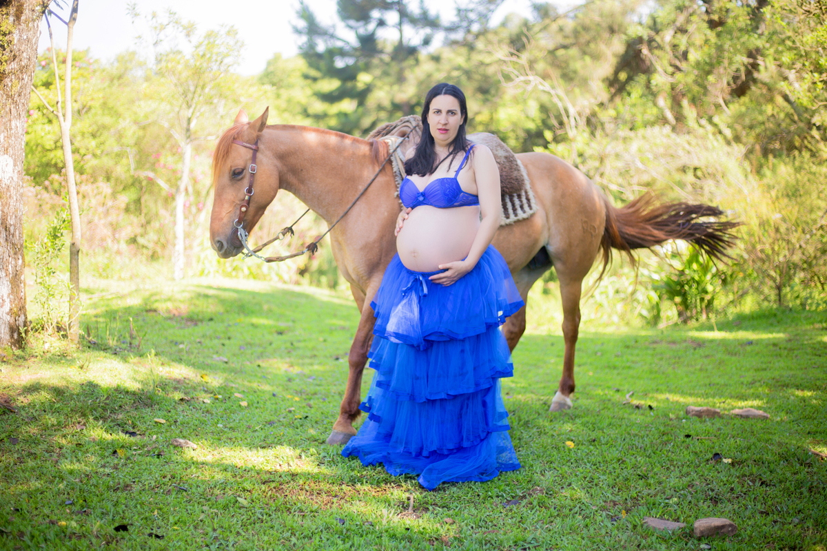 a gestante segura a rédea de cavalo enquanto posa para a fotógrafa em sua sessão fotográfica