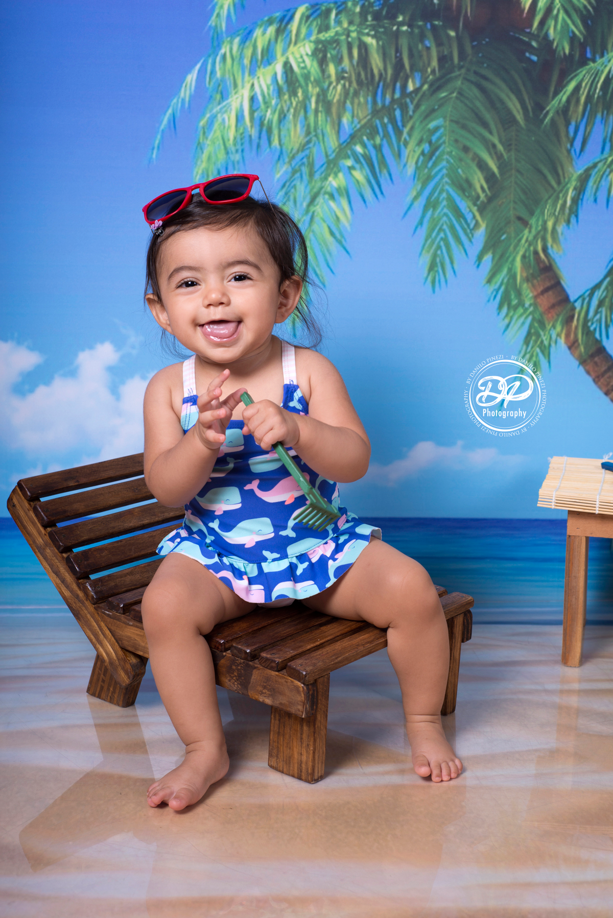 Sessão fotográfica de bebê menina na praia, com óculos e espreguiçadeira