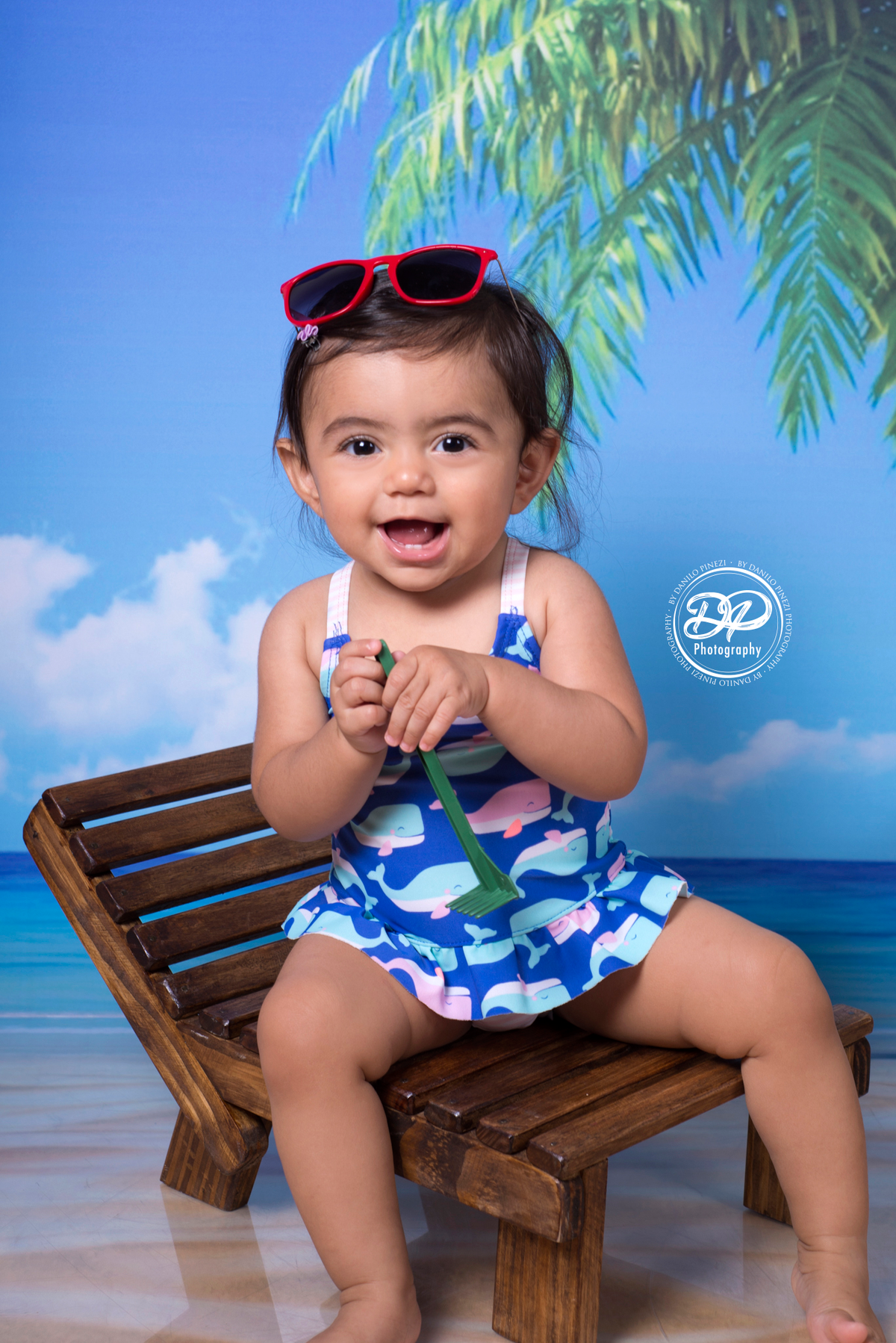 Sessão fotográfica de bebê menina na praia, com óculos e espreguiçadeira