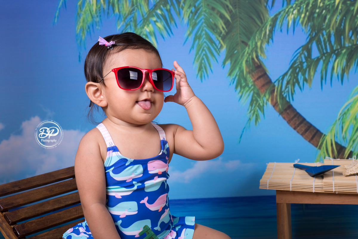 Sessão fotográfica de bebê menina na praia, com óculos e espreguiçadeira