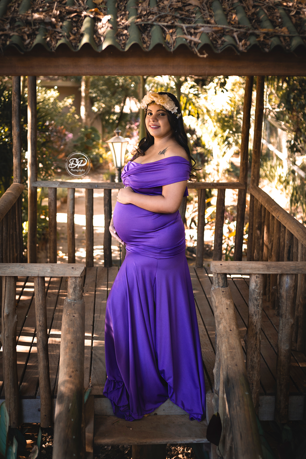 gestante com vestido roxo em pé, de perfil com as mãos na barriga, em uma casinha na árvore