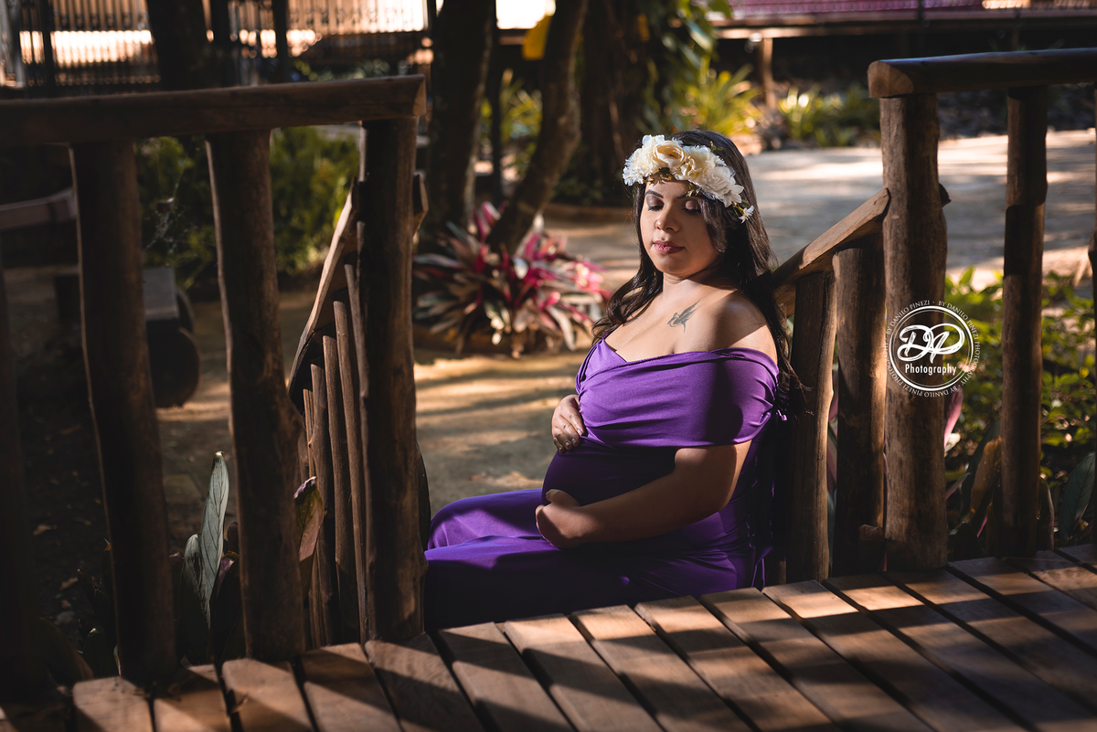 gestante com vestido sentada nos degraus de uma escada, com as mãos na barriga usando uma coroa de flores.