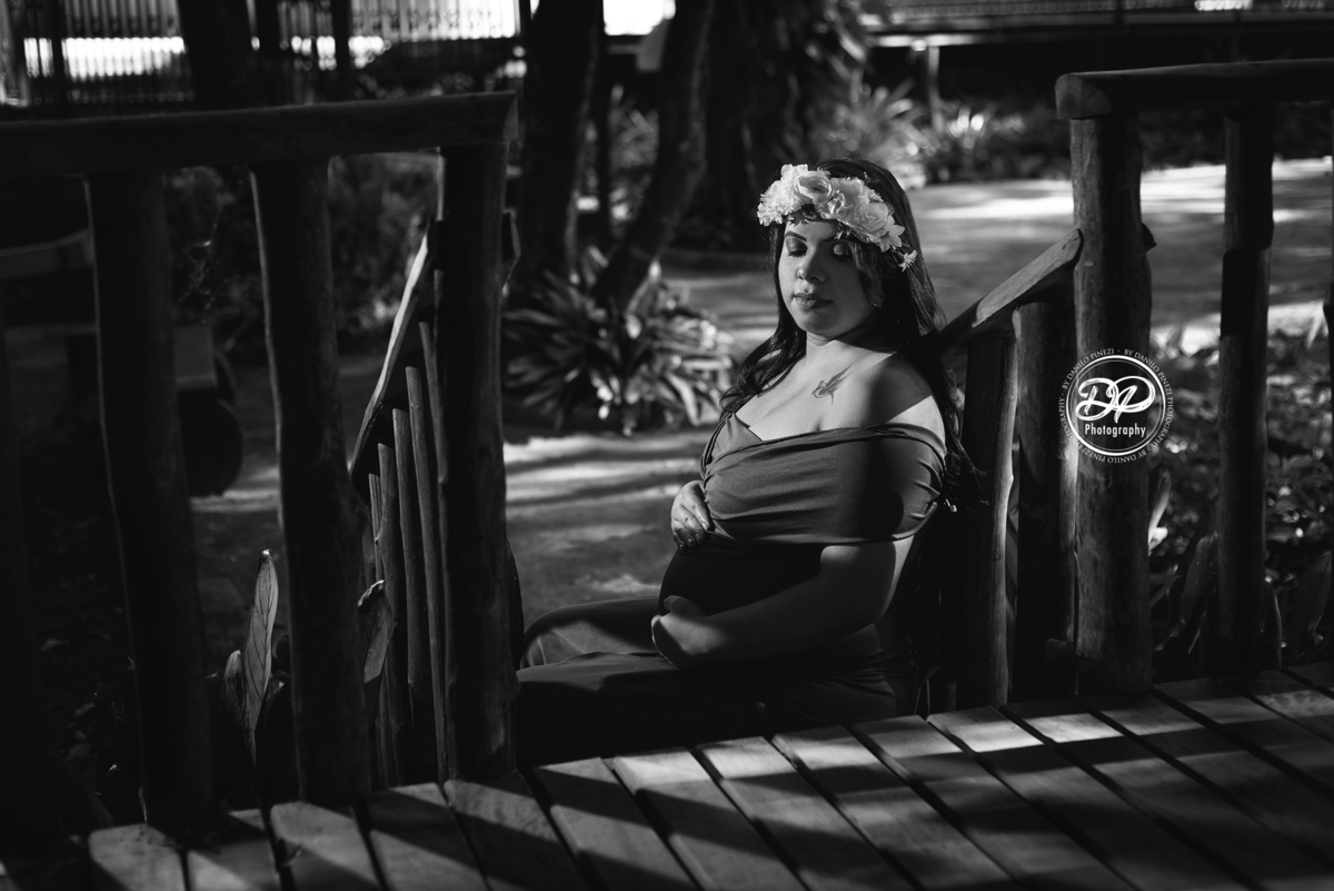 gestante com vestido sentada nos degraus de uma escada, com as mãos na barriga usando uma coroa de flores. Fotografia gestante preto e branco