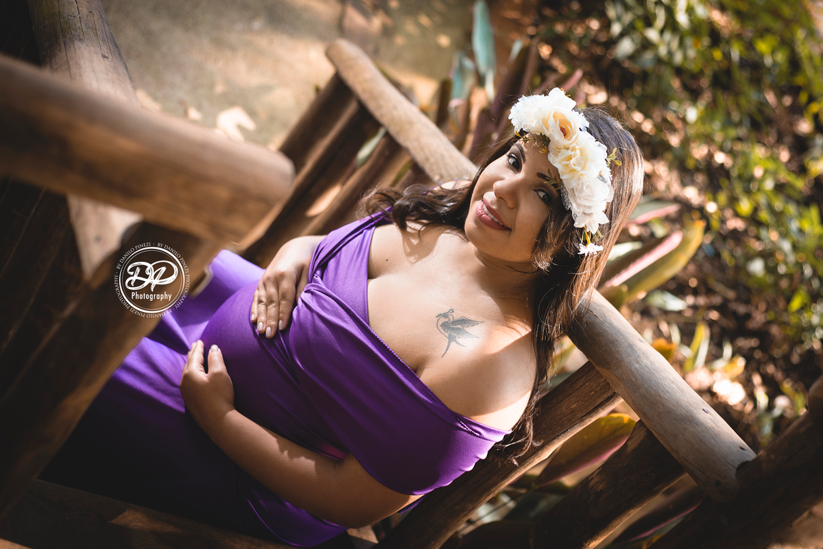 Gestante com vestido sentada nos degraus de uma escada, com as mãos na barriga usando uma coroa de flores. 