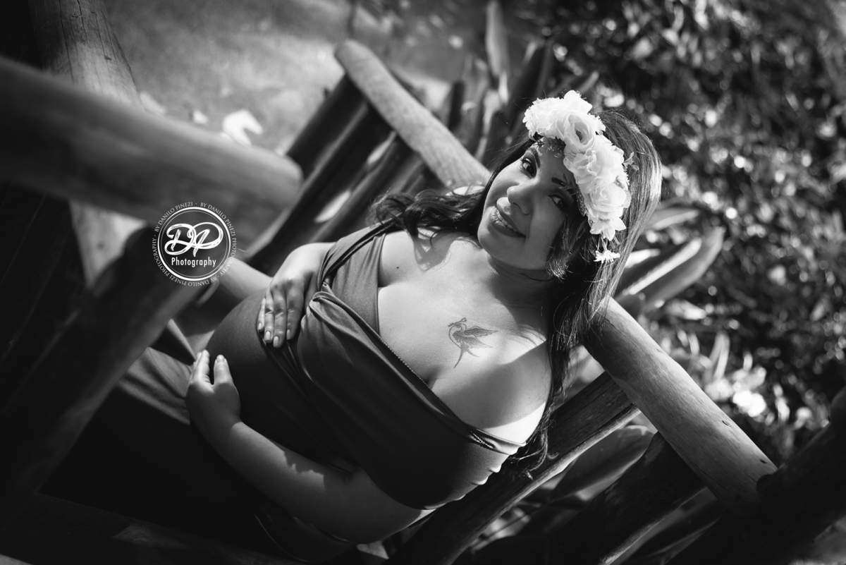 gestante com vestido sentada nos degraus de uma escada, com as mãos na barriga usando uma coroa de flores. Fotografia gestante preto e branco