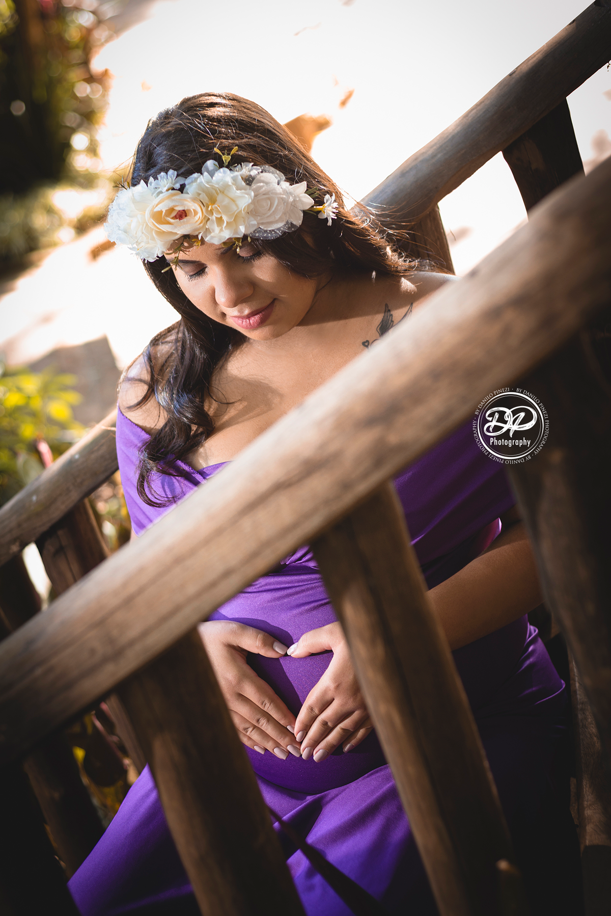 Gestante com vestido sentada nos degraus de uma escada, com as mãos na barriga usando uma coroa de flores.  Gestante fazendo um coração com as mãos sobre a barriga