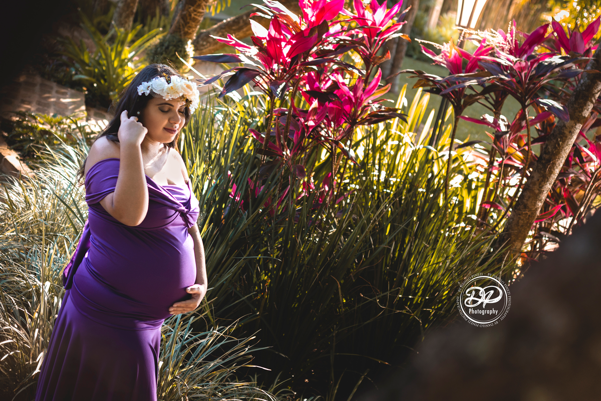 gestante com vestido roxo em pé, de perfil com uma das mãos na barriga e a outra arrumando o cabelo,  