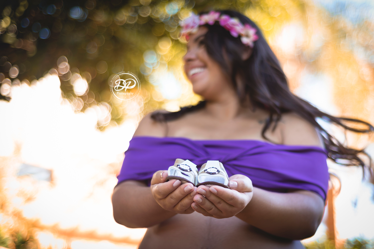 gestante com vestido roxo em pé, segurando nas mãos os sapatinhos de seu bebê. 