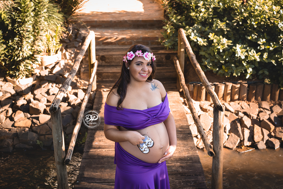 gestante com vestido roxo em pé sobre uma ponte de madeira,  com uma das mãos na barriga e a outra segurando os sapatinhos de seu bebê