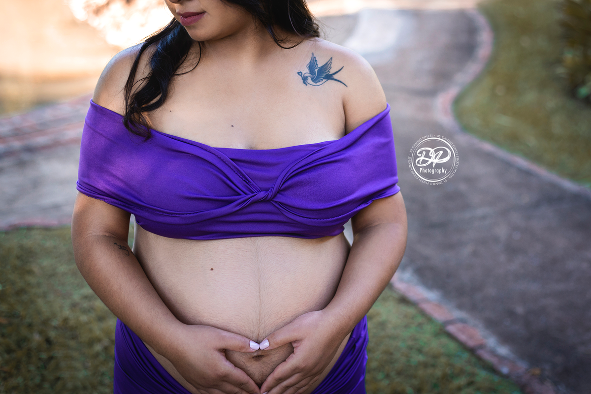gestante com vestido roxo em plano americano, mostrando sua tatuagem de beija flor, fazendo um coração com as mãos sobre a barriga