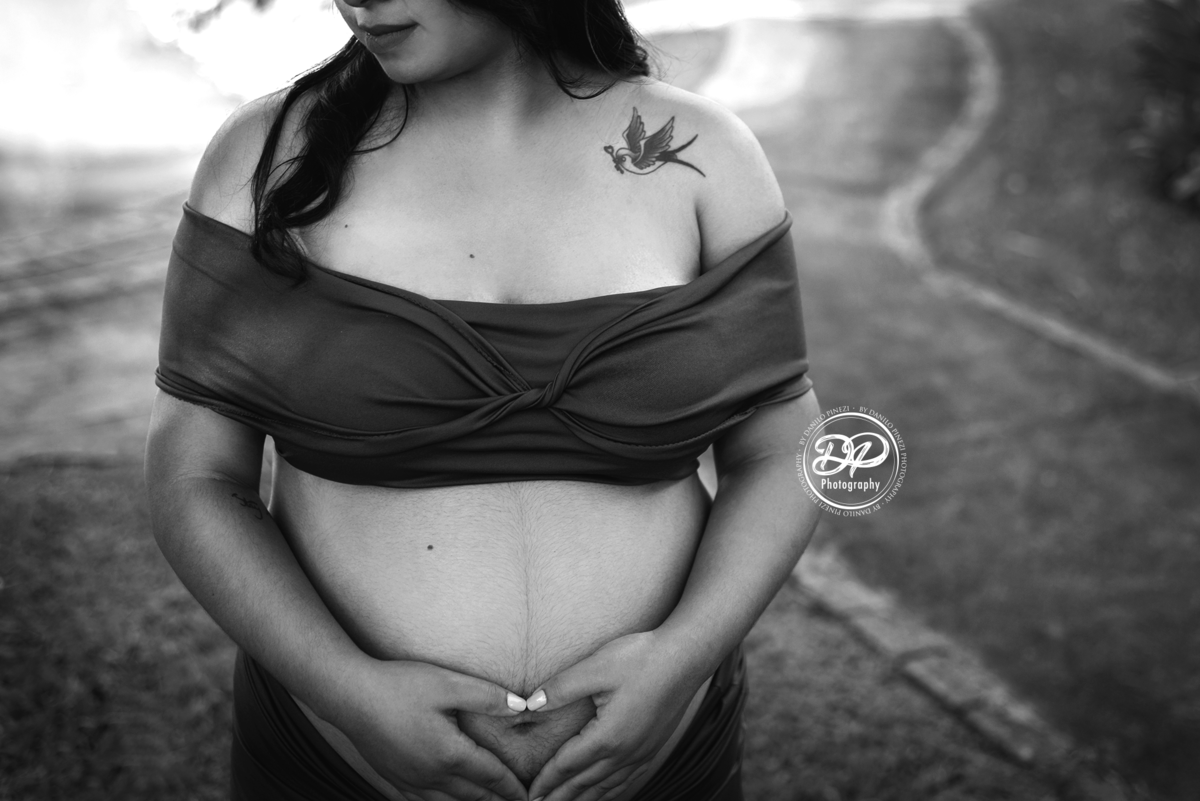 gestante com vestido roxo em plano americano, mostrando sua tatuagem de beija flor, fazendo um coração com as mãos sobre a barriga. Fotografia de gestante em preto e branco