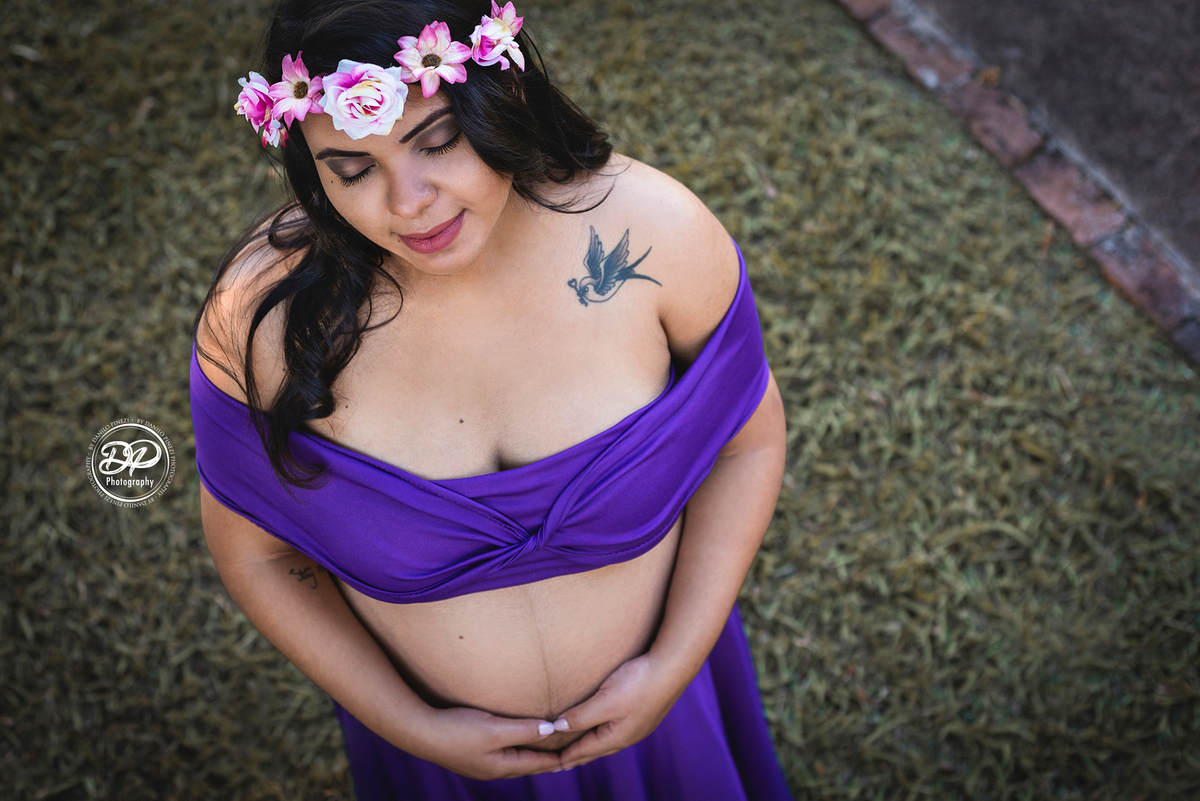 gestante com vestido roxo e coroa de flores, em plano americano, mostrando sua tatuagem de beija flor, fazendo um coração com as mãos sobre a barriga
