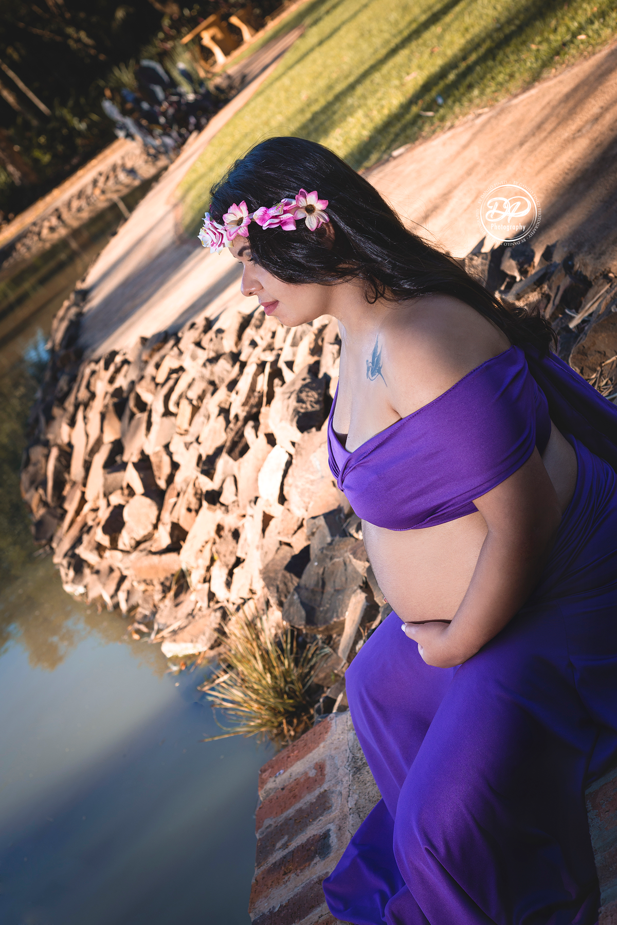 Gestante com vestido roxo, sentada a beira de um lago, com as mãos na barriga e olhando para o horizonte