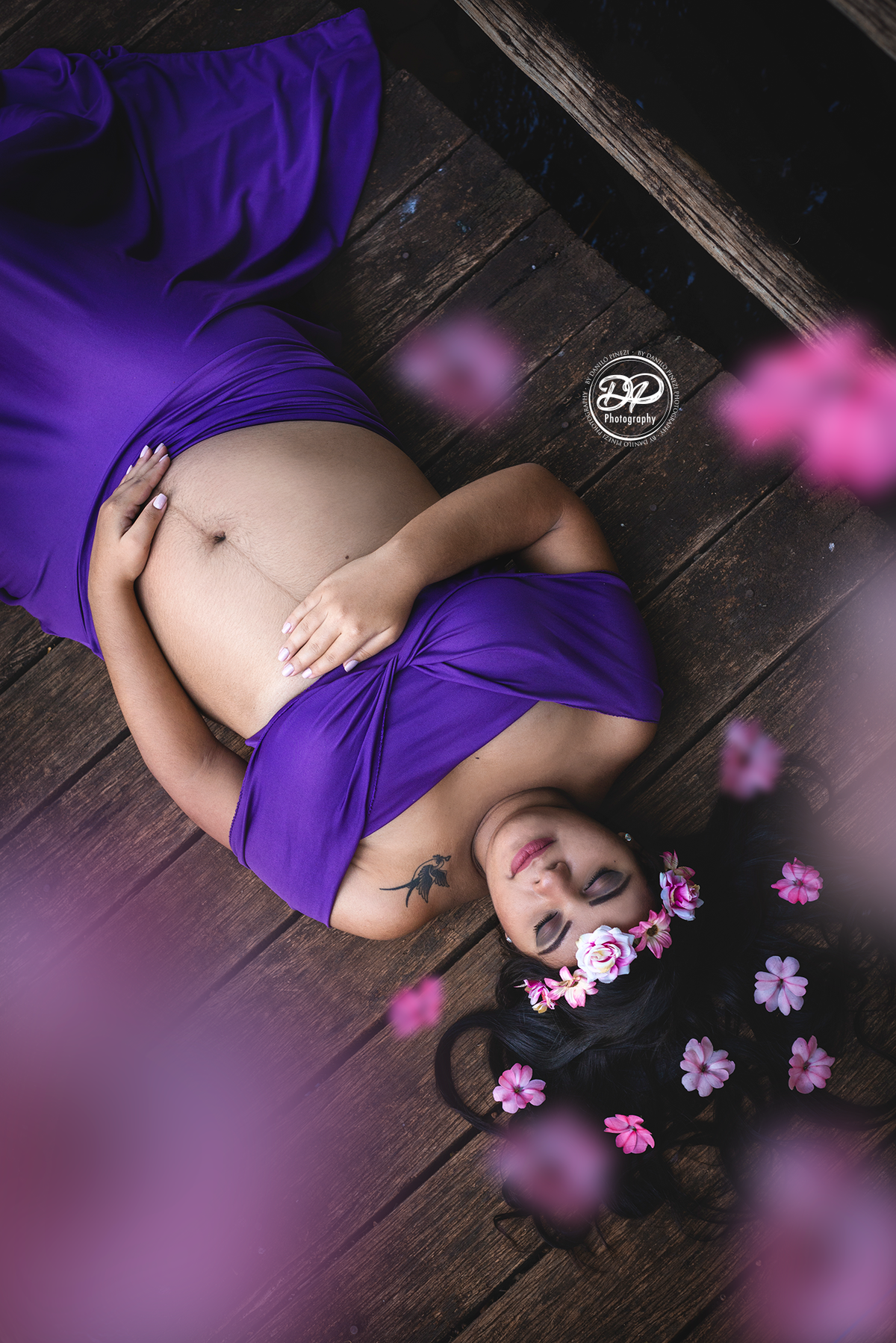 Gestante com vestido roxo, deitada sobre uma ponte de madeira, com as mãos na barriga, e uma chuva de flores caindo sobre si.