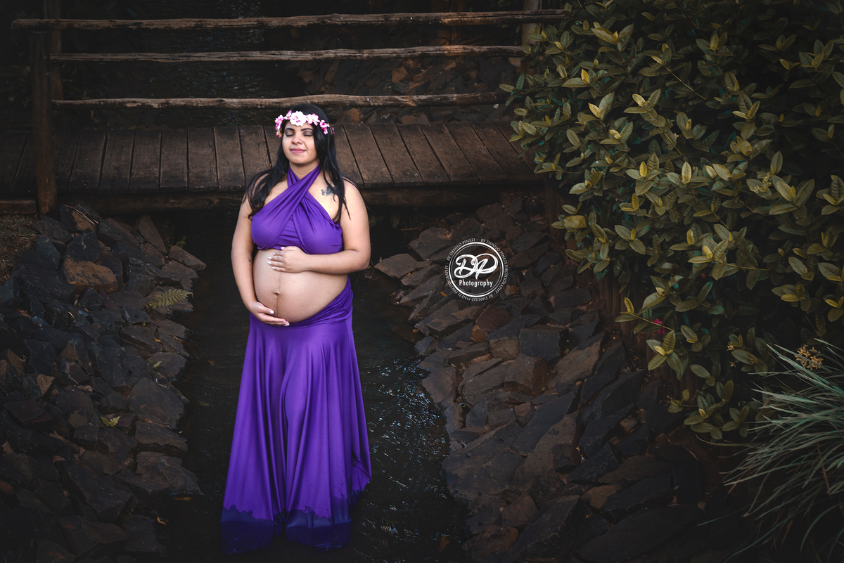 Gestante com vestido roxo, caminhando dentro um pequeno córrego com as mãos na barriga 