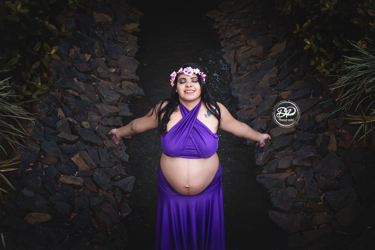 Gestante com vestido roxo, caminhando dentro um pequeno córrego com os braços abertos sentindo o vento bater em seu rosto