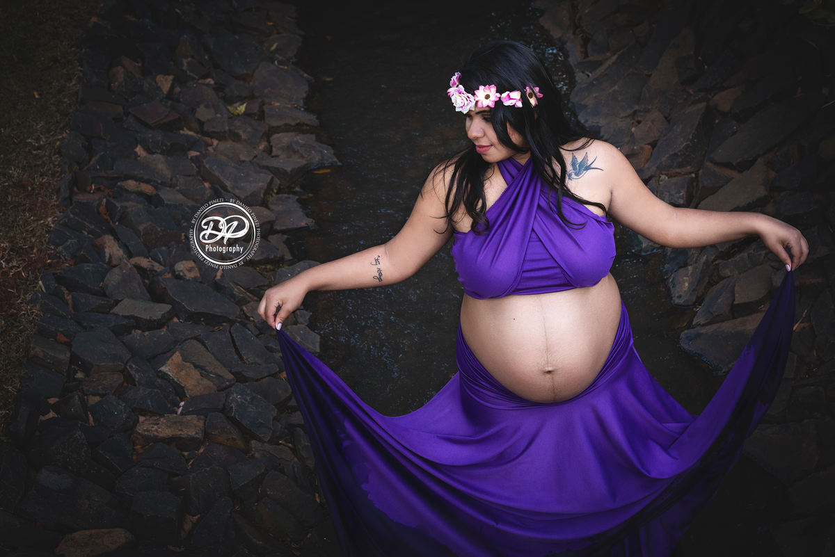 Gestante com vestido roxo, caminhando dentro um pequeno córrego segurando as pontas de seu vestido