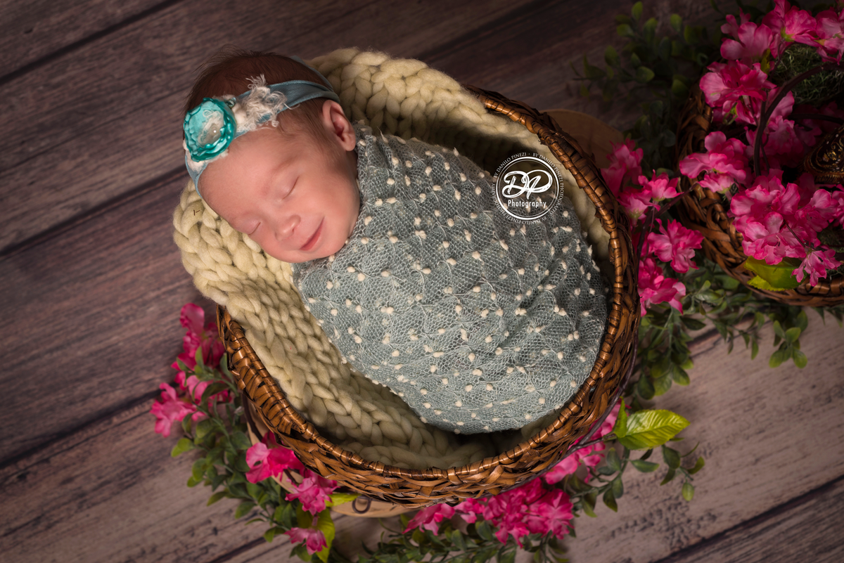 Bebê recém nascida sorrindo, dormindo tranquilamente enrolada em wrap pipoca, dentro de um cesto enfeitado com cerejeiras,  em sessão newborn realizada no Estúdio Danilo Pinezi Fotografia em Bariri