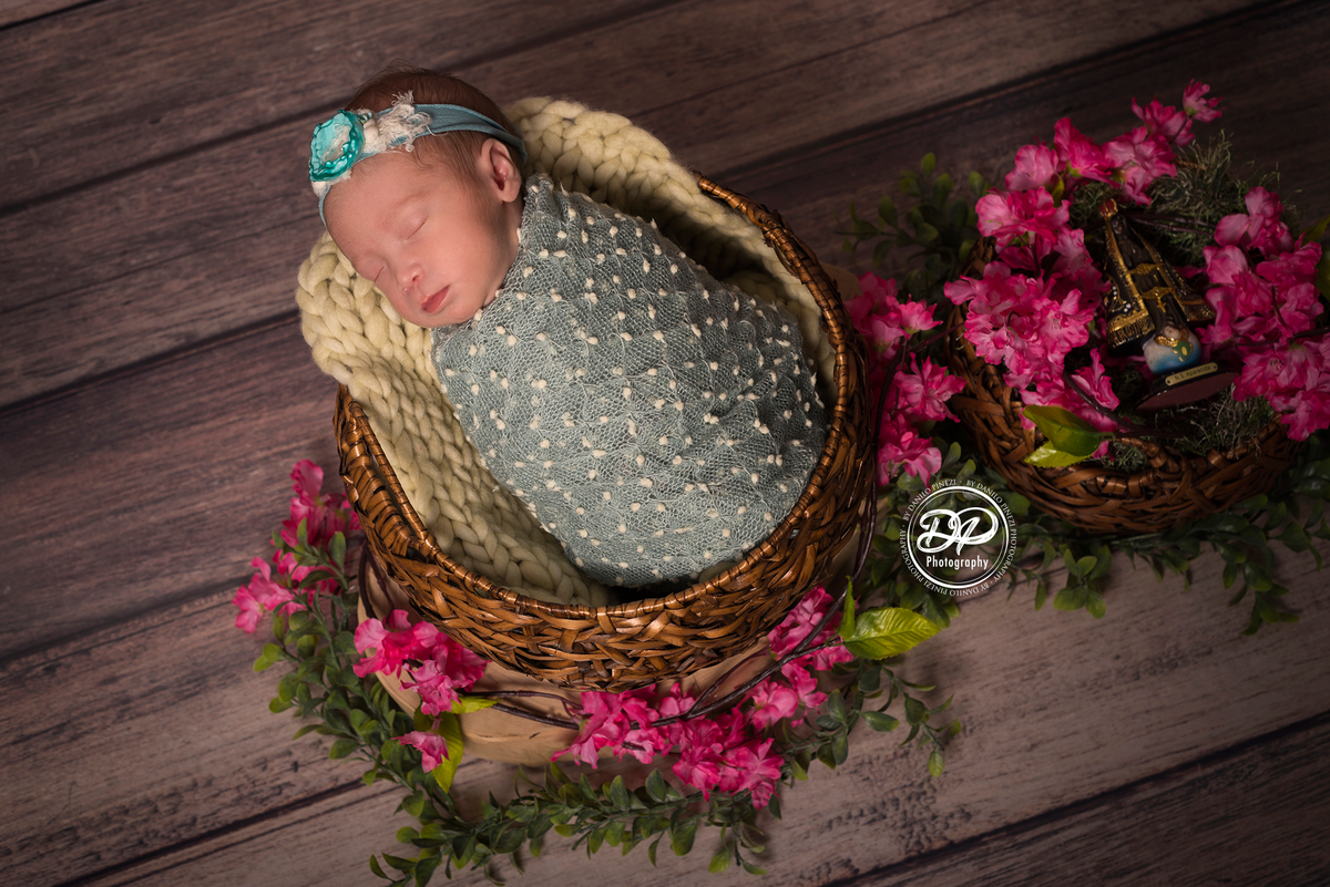 Bebê recém nascida sorrindo, dormindo tranquilamente enrolada em wrap pipoca, dentro de um cesto enfeitado com cerejeiras e uma imagem de Nossa Senhora Aparecida,  em sessão newborn realizada no Estúdio Danilo Pinezi Fotografia em Bariri