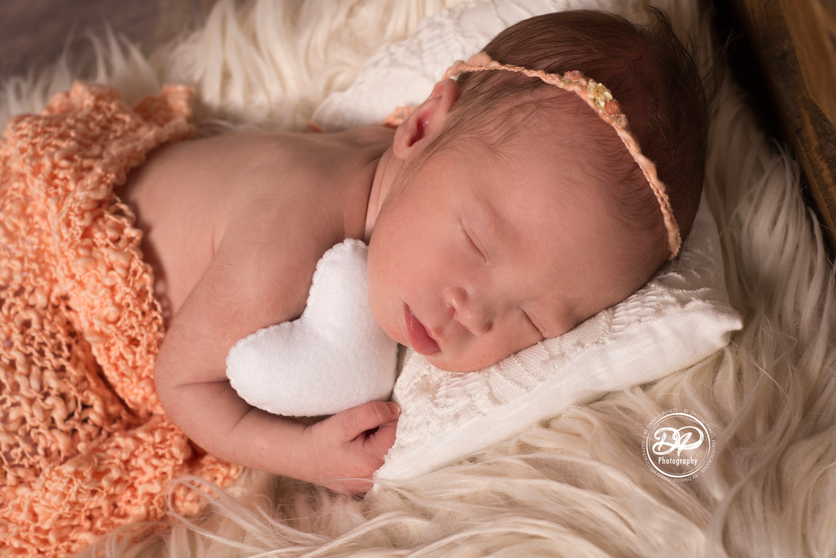 Bebê recém nascida segurando um coração, deitada em uma caminha, dormindo tranquilamente em sessão newborn realizada no Estúdio Danilo Pinezi Fotografia em Bariri