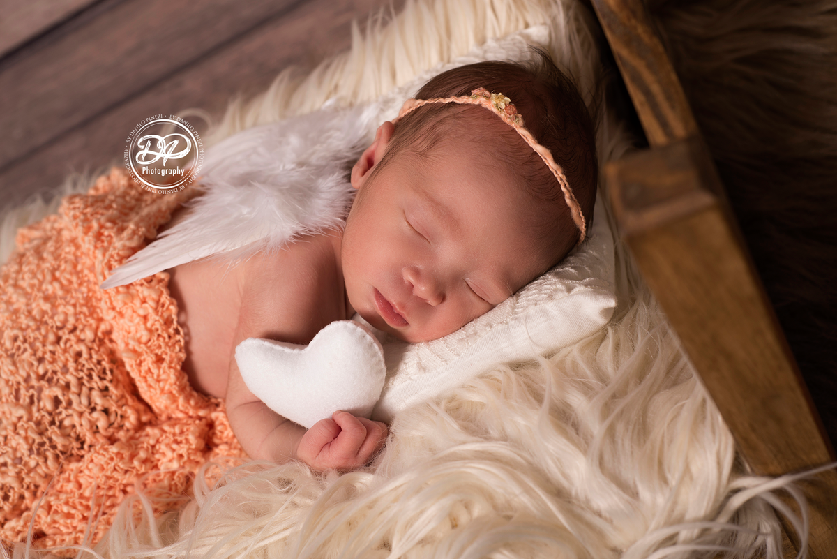Bebê recém nascida vestida de anjo, deitada em uma caminha segurando um coração, dormindo tranquilamente em sessão newborn realizada no Estúdio Danilo Pinezi Fotografia em Bariri
