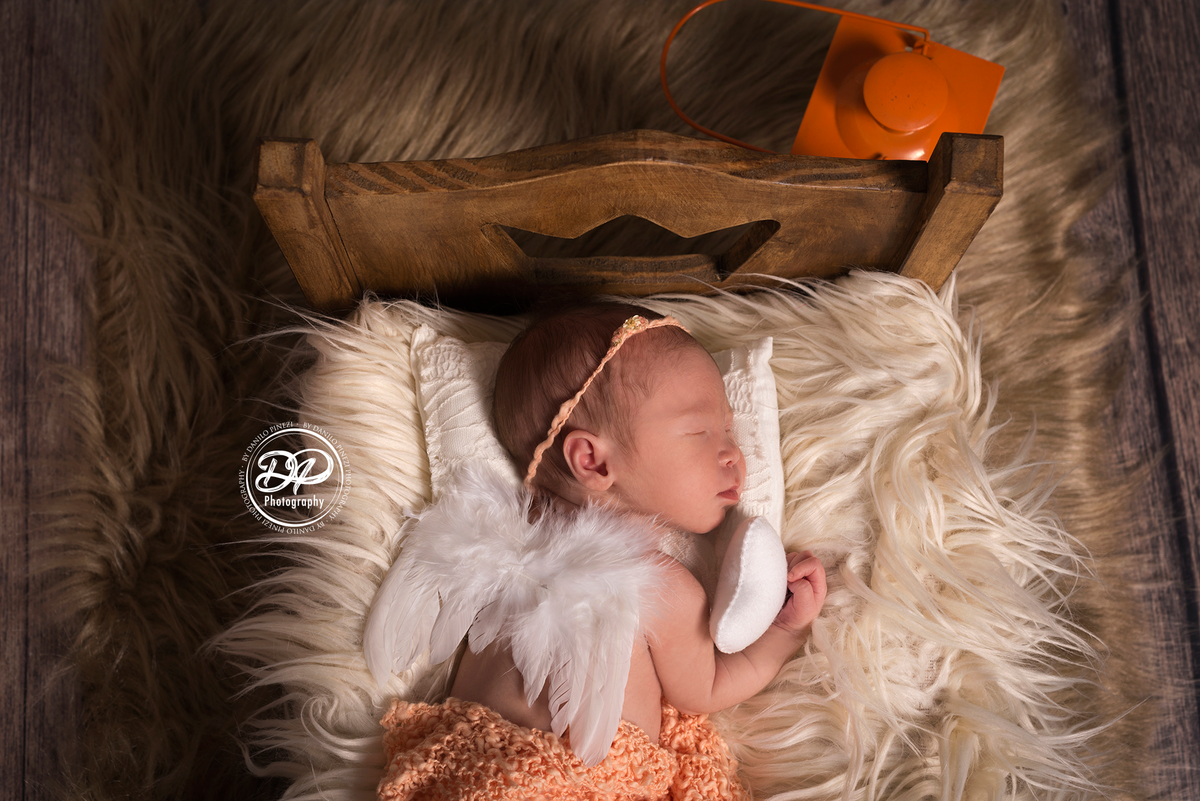 Bebê recém nascida vestida de anjo, deitada em uma caminha, dormindo tranquilamente em sessão newborn realizada no Estúdio Danilo Pinezi Fotografia em Bariri