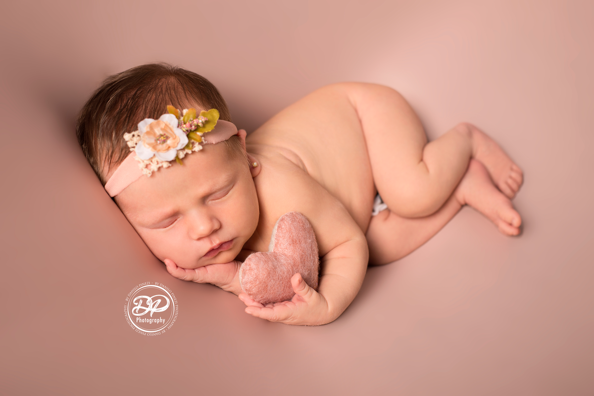 Newborn de menina executado em Bariri, pose de ladinho segurando um coração no fundo rosé, estúdio Danilo Pinezi Fotografia