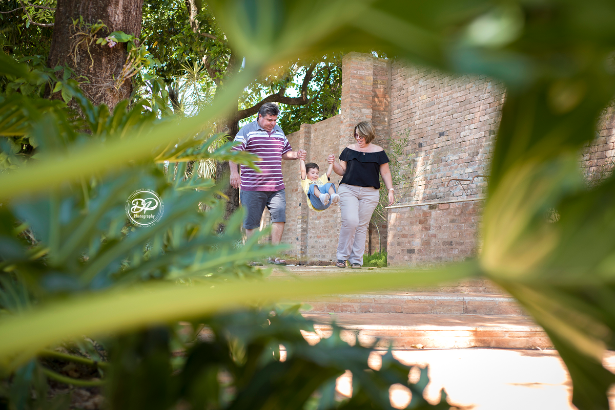 Avós andando com netinho saltando feliz em um jardim, em ensaio família  externo feito em bariri/ SP.