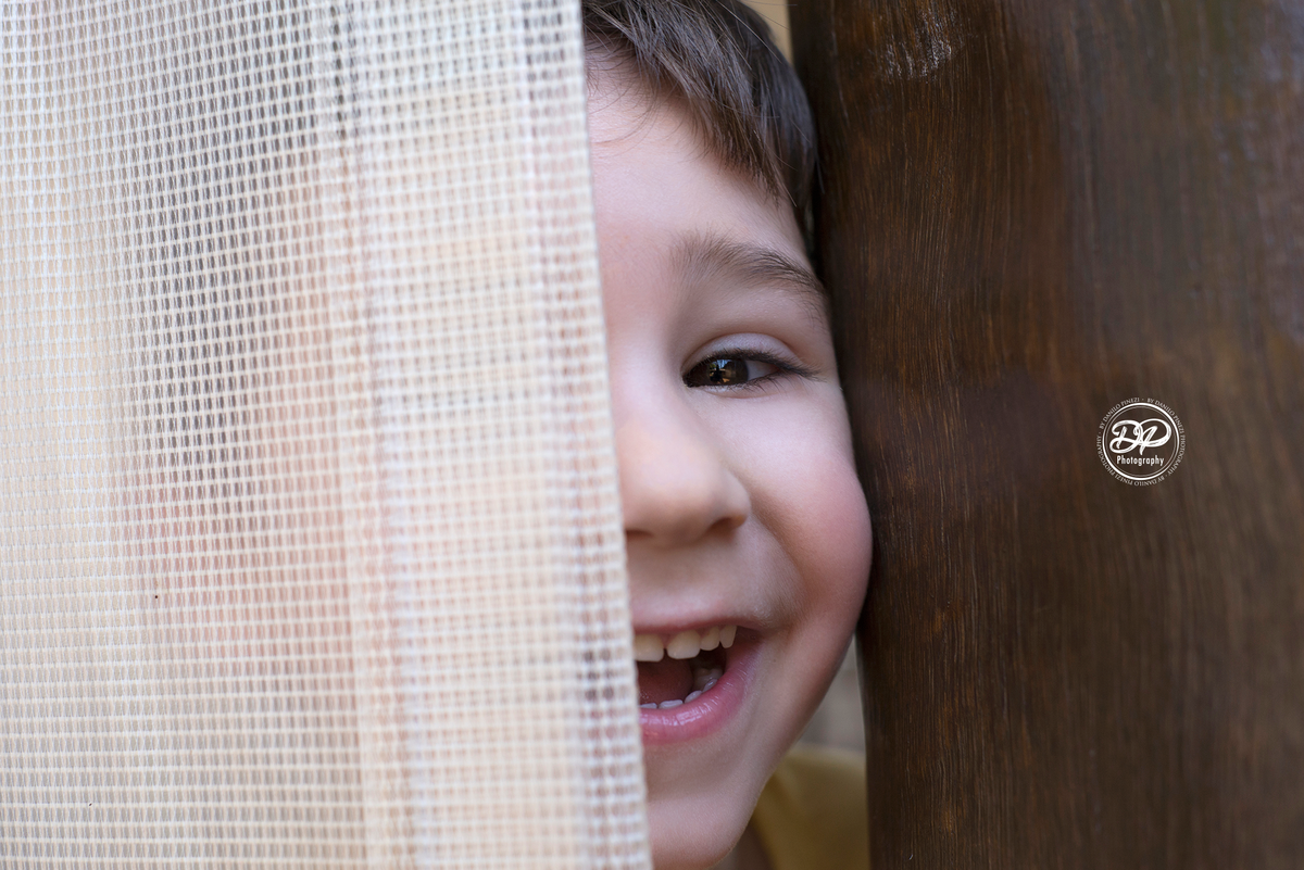 menino sorrindo, com meio rosto aparecendo, brincando de pique esconde,  em ensaio família realizado em Bariri/SP